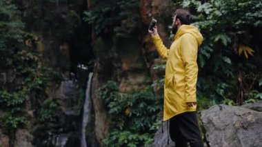 Sarı ceketli bir adam elinde akıllı telefonu tutar ve şelale ve güzel doğanın videosunu çeker. Seyahatte cep telefonu kamerasıyla şelale çeken genç erkek turist.