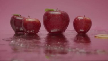 Pembe bir yüzeyde üç kırmızı elma sergileniyor, biri suda, biri de bir bardak suda. Video, elmaların suyla etkileşimini vurguluyor. Doğal ve çekirdeksiz meyveler olarak gösteriliyor.