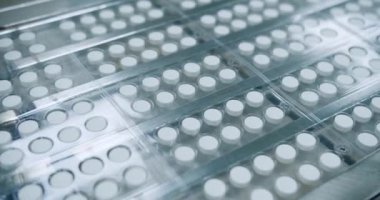 Bir fabrikadaki konveyör bandında beyaz hapların üretimini gösteren bir video. Plastik kaplarda ve eczanelerde yakın plan haplar ve tepsiler var.