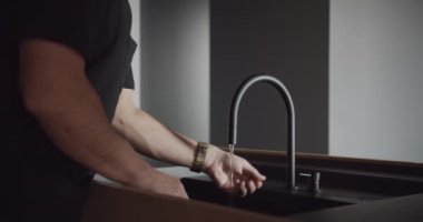 Bir adam mutfak lavabosunda ellerini yıkıyor, musluğu açmak için parmağını kullanıyor. Döşeme sert ahşaptan yapılmış, bileşik bir malzeme.