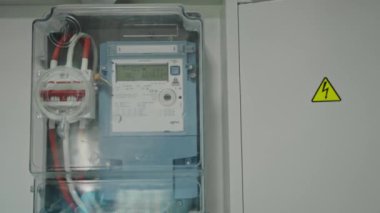Uyarı işaretinin yanındaki şeffaf bir kutuda bulunan gelişmiş bir elektrik sayacının net görüntüsü. Ofis Duvarında Şeffaf Kutuda Modern Elektrik Taksimetresi