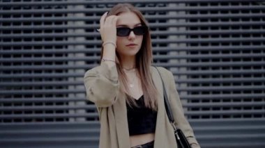 Güneş gözlüklü şık kıyafetli modaya uygun genç bir kadın şehirde modern bir metal şebekeye karşı güven içinde poz veriyor. Güneş Gözlüklü Şık Genç Kadın Şehir Ayarlarında Pozisyon