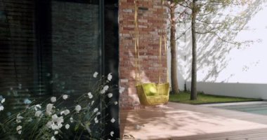 Çağdaş bir evin dış görünüşünün davetkar bir görüntüsü. Stili bir hasır asılı sandalye, sakin bir bahçe ortamı ile tamamlanmış. Wicker Asma Sandalyesi ve Lush Garden ile Modern Ev Dış Görünümü