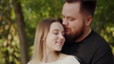 Genç bir erkek ve kadın kucaklaşıyor, gözleri kapalı, yemyeşil bir parkta sıcak güneşin altında huzurlu bir anın tadını çıkarıyorlar. Sunny Park 'ta İhale Anı Paylaşan Genç Çift