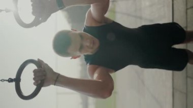 Spor salonunda jimnastik halkaları kullanan odaklanmış genç bir erkek atlet hafif ışıklandırma altında güç ve konsantrasyon gösteriyor. Genç Erkek Sporcu Jimnastik Yüzükleri Egzersizi Salonu 'nda. Dikey video.