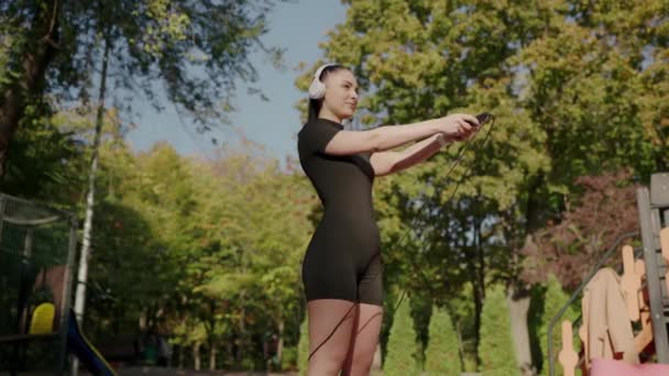黒いアクティブなスーツの若い女性が公園で暖かい秋の日を楽しんで 白いヘッドフォンを通して音楽を聴いています 晴れた秋の日に公園でストレッチする若い女性 ロイヤリティフリーストック映像