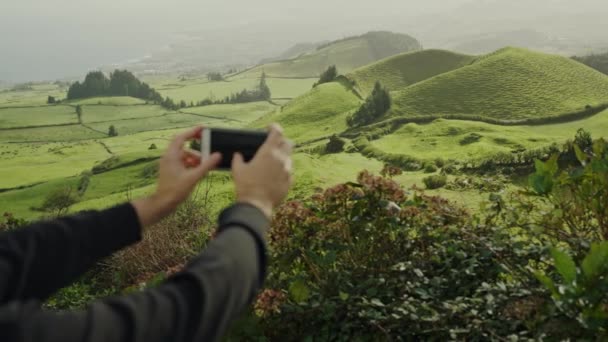 若い男はスマートフォンを持ち 美しい緑の丘のビデオを作ります 男性は携帯電話のカメラを使用して谷で風景の写真を撮ります 動画クリップ