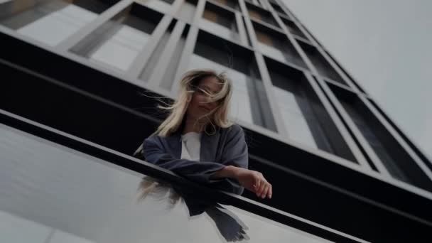 一个穿着休闲装的年轻女人靠在栏杆上 凝视着 身后是一座高耸的现代建筑 年轻女子靠铁路反对现代建筑 — 图库视频影像