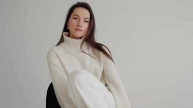 Beyaz kazaklı, pantolonlu, sandalyede oturan ve stüdyoda poz veren güzel esmer kadın. Güzel bir kadının pürüzsüz ve sağlıklı bir teni var. Kameraya bakan muhteşem kadının portresi..