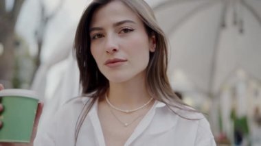 Elinde kahve fincanı tutan genç bir kadının portresi, beyaz bir bluz giymiş, kentsel bir ortamda, yumuşak odaklı arka planda. Genç Kadın Şehir Düzeninde Kahve Keyfini Çıkartıyor, Gelişigüzel Tarz