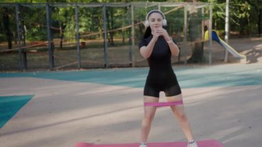 Kulaklıklarla donatılmış açık hava parkı ortamında direniş bandıyla çömelme egzersizleri yapan kadın sporcu. Park 'ta Direniş Bandosu ile Açık havada Atletik Kadın Eğitimi