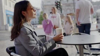 Profesyonel kıyafetli genç bir kadın, şehirde insanlar geçerken elinde kahve fincanı olan açık bir kafede huzurlu bir anın tadını çıkarıyor. Genç Profesyonel Kadın Şehirde Açık Cafe 'de Dinleniyor