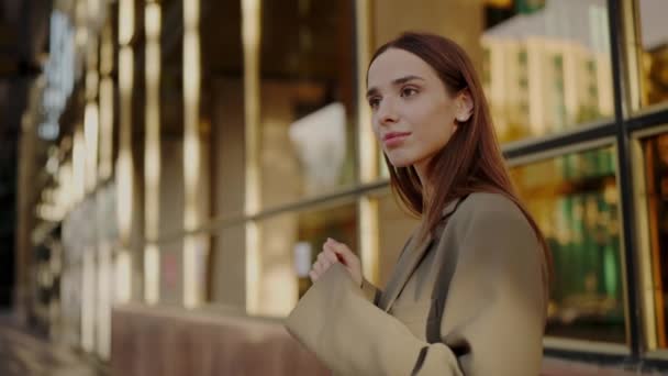 一位穿着时髦风衣的优雅的年轻女子站在一座现代玻璃建筑前 映衬着金色的落日 夕阳西下城市环境中自信的年轻女性 — 图库视频影像