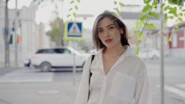 Beyaz bluzlu şık bir genç kadın bir şehir caddesinde duruyor, gözlerini kapıyor ve yavaşça kameraya bakıyor. Şık Genç Kadın Alacakaranlıkta Şehir Caddesinde Poz Veriyor