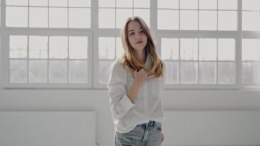 Sakin, beyaz bir gömlek ve kot giymiş genç bir kadın, parlak, havadar bir odada pencerenin önünde düşünceli bir şekilde poz veriyor. Beyaz Gömlekli Genç Kadın Düşünceli Poz veriyor Pencere kenarında