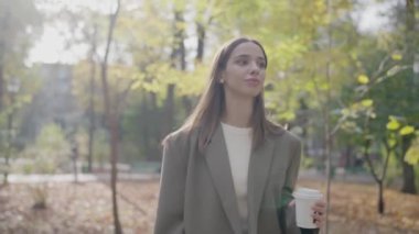 Elinde kahve fincanı olan genç profesyonel bir kadın, sonbaharda güneşli bir şehir parkında huzurlu bir şekilde yürüyor ve sakin bir şehir yaşam tarzı sergiliyor. Genç Kadın Şehir Parkı 'nda Huzurlu Sonbahar Gününün keyfini çıkarıyor