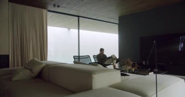 Modern stil oturma odasında rahat bir şekilde televizyon seyreden bir adamın sakin bir sahnesi. Minimalist iç tasarım. Modern Oturma Odasında Sandalyede Rahatlayan Adam