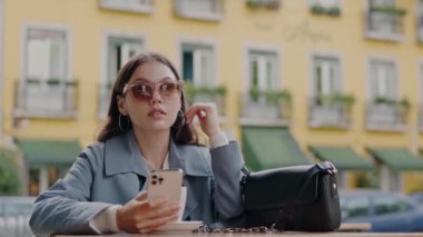Trençkotlu neşeli bir kadın akıllı telefonunu kullanıyor, renkli şehir binalarının önünde taksi için el sallıyor. Şehirde Taksi Çalarken Akıllı Telefon Kullanan Gülümseyen Kadın