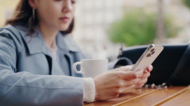 Mavi ceketli odaklanmış genç bir kadın açık bir kafe masasında akıllı telefonunu kullanarak şehirdeki modern hayatın bir anını yakalıyor. Cafe Table 'da akıllı telefon kullanan genç bir kadın.