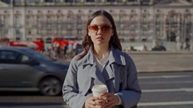 Şık genç bir kadın güneşli bir şehirde elinde kahveyle dolaşıyor. Modayı takip eden güneş gözlüğü ve modern bir trençkot giyiyor. Şık Genç Kadın Şehirde Yürüyor
