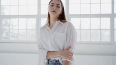 Gevşek beyaz bir gömlek ve kot pantolon giyen zarif genç bir kadın büyük pencereli, güneşli bir stüdyoda güvenle duruyor. Beyaz Gömlekli Genç Kadın Parlak Stüdyoda Gelişigüzel Poz Veriyor