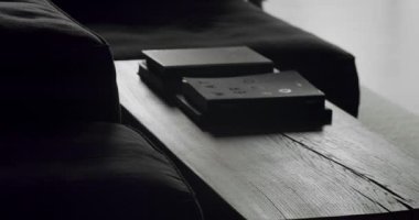 Modern bir Minimalist oturma odasında, siyah mobilyalar, ahşap masa ve kitapların olduğu yerleri kapatın. Adam masanın yanında yürür ve siluetler ve gölgeler yaratır..