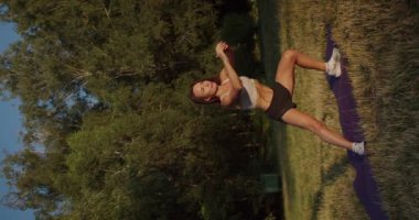 Atletik kadın sabah antrenmanına çıkıyor ve paspasın üzerinde yan hamleler yapıyor. Dişi sporcu kasları ısıtıp esnetiyor. Atletik sarışın kasları eğitir. Dikey video.