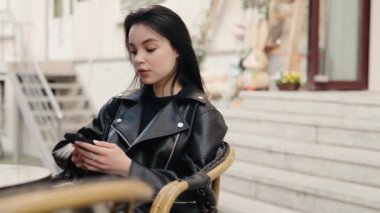 A young woman in a black leather jacket sits outdoors, holding and using her mobile phone. The image conveys a sense of casual and modern urban lifestyle. Young Woman Using Mobile Phone