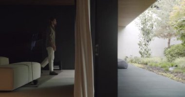 A male adult stands contemplating in a modern, well-lit house, gazing peacefully into a lush garden through transparent glass walls. Man Reflecting in Modern Home With Garden View Through Glass