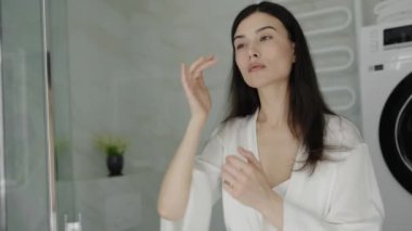 Beyaz bornozlu bir kadın modern, parlak bir banyoda cilt bakımını yapıyor. Sahne, sakin ve öz-bakım hissi veriyor. Banyo Aynası Yansıması 'na Cilt Rutini Uygulayan Kadın