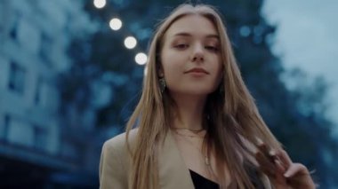 Şık giyimli genç bir kadın alacakaranlıkta bir şehir caddesinde duruyor. Şehrin ışıkları yumuşak bir bokeh arkaplan oluşturuyor. Şık Genç Kadın Alacakaranlıkta Sokak Işıklarıyla Bir Şehirde Duruyor