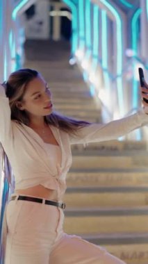 Geceleri ışıl ışıl yanan bir şehir merdiveninde dururken selfie çeken gülümseyen bir kadın. Mutlu görünüyor ve günlük beyaz kıyafetler giyiyor. Geceleyin Renkli Aydınlık Şehir 'de Selfie çeken Kadın