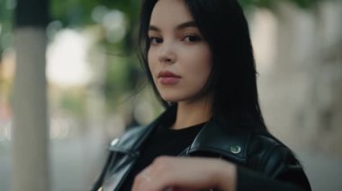 Siyah deri ceket giyen, kendinden emin ve ciddi görünen genç bir kadının açık hava portresi. Bulanık arkaplan onun güçlü ifadesini ve tarzını vurguluyor..