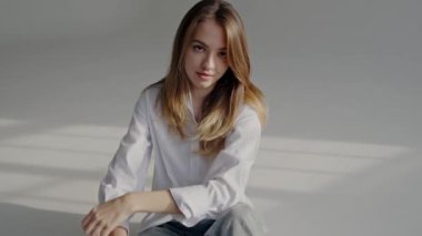 Beyaz tişört giyen sakin genç bir kadının portresi, hafif ışıklandırılmış bir stüdyoda düşünceli bir ifadeyle gündelik bir şekilde oturuyor. Serene Genç Kadın Beyaz Gömlekli Stüdyoda Gelişigüzel poz veriyor