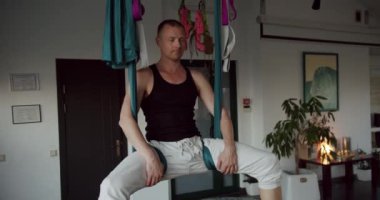 Handheld shot of strong male hanging upside down on aerial hammock and bending body during yoga session in light studio. Working out in class, performing aero yoga. Yoga class together indoors.
