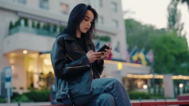 Siyah deri ceketli genç bir kadın şehir ortamında akıllı telefonuna dalmış bir şekilde dışarıda oturuyor. Arkaplanda bayraklar ve kozmopolit bir ortam izlenimi veren modern bir bina yer alıyor..