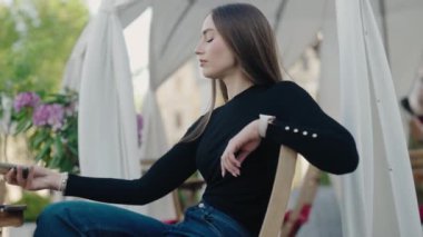 Gündelik giysiler içinde genç bir kadın dışarıda oturmuş, akıllı telefonunu dikkatle izliyor. Arka plan yeşillik ve beyaz kumaş yapılarından oluşuyor. Genç kadın dışarıda dinleniyor.