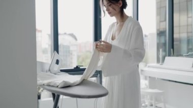 Beyaz elbiseli genç bir kadın geniş pencereli, parlak, modern bir dairede giysilerini ütülüyor. Huzurlu ve temiz bir ortamda aile içi aktiviteler. Parlak Daire 'de Giysileri Ütüleyen Kadın