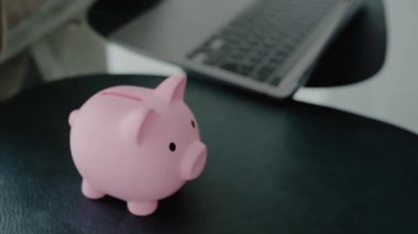 Birikimleri ve finansal planlamaları sembolize eden, arka planda dizüstü bilgisayarı olan pembe domuz kumbarasına yazı-tura atan bir el. Arka planda dizüstü bilgisayarı olan Pembe Piggy Bank 'a madeni para sokmak.