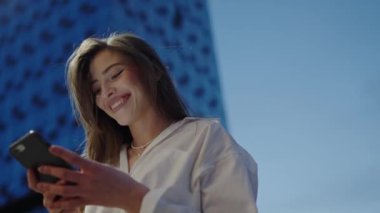 Genç bir kadın, arka planda modern mavi bir binayla dışarıda akıllı telefonuna bakıp gülümsemeyi seviyor. Şehir Ayarı Günışığında Akıllı Telefon Kullanan Gülümseyen Kadın