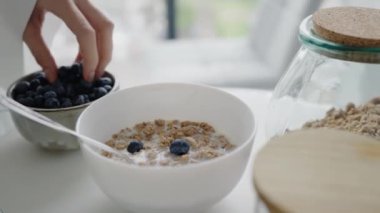 Sağlıklı bir kahvaltı için bir kase granola ve süte taze yaban mersini ekleyen bir elin yakın çekimi. Yakınlarda bir kavanoz granola ve bir kase yaban mersini var. Kahvaltıya Yabanmersini Ekleme Elleri