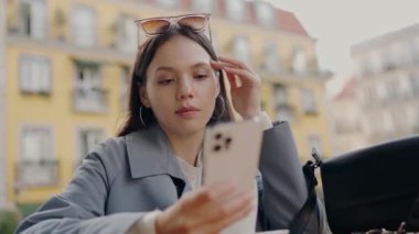 Tarz sahibi gri ceketli odaklanmış genç bir kadın arka planda renkli binalar bulunan hareketli bir şehir caddesinde akıllı telefonunu kullanıyor. Şehir Caddesinde Akıllı Telefon Kullanan Genç Kadın, Gelişigüzel Tarz