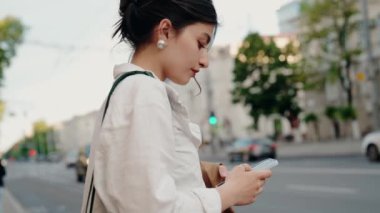 Bir yaz günü şehir caddesinde yürürken akıllı telefonunu kullanan genç bir kadının yan görüntüsü. Arka planda arabalar ve binalar var, şehir ortamını gösteriyor. Akıllı Telefon Kullanan Kadın