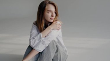 Beyaz gömlekli beyaz bir kadın minimalist bir stüdyo ortamında otururken düşünceli görünüyor. Sakin ve içsel bir ruh hali sergiliyor. Stüdyoda Düşünceli İfadesi Olan Zarif Genç Kadın.