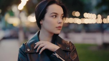 Siyah deri ceketli zarif bir kadın arka planda bulanık şehir ışıklarıyla düşünceli bir şekilde uzaklığa bakıyor. Deri ceketli Genç Kadın Portresi Arka planda Neşeli Işıklar