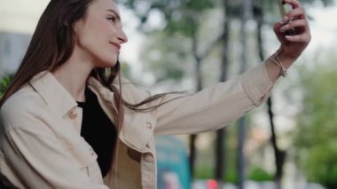 Uzun saçlı genç bir kadın güneşli bir günde açık hava parkında akıllı telefonuyla selfie çekiyor. Güneşli bir günde parkta selfie çeken genç bir kadın.