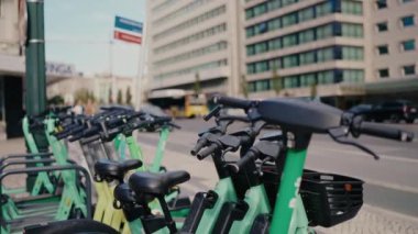 Şehir caddesinde park etmiş çok sayıda yeşil elektrikli scooter modern ofis binalarının yakınında kentsel ulaşım seçenekleri sunuyor. Kalabalık bir şehir ortamında işe gidip gelmek için sürdürülebilir bir seçim.