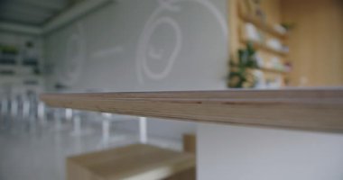 Modern bir kahve dükkanındaki ahşap bir masanın odaklanmış görüntüsü. Bulanık arka plan raflar ve dekoratif bitkilerle şık bir minimalist iç mekan ortaya çıkarıyor, sakin ve misafirperver bir atmosfer yaratıyor..