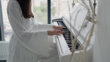 Beyaz cüppeli bir kadın, şehre tepeden bakan büyük pencereli parlak bir odada piyano çalıyor. Beyaz cüppeli, Parlak Odada Piyano Çalan Kadın