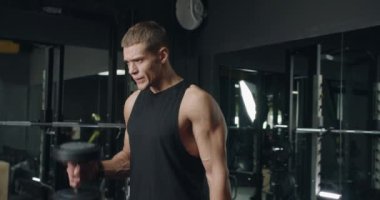 Kolsuz tişörtlü kararlı genç bir erkek yoğun bir şekilde dambıl kaldırıyor, gücünü gösteriyor ve iyi donanımlı bir spor salonuna odaklanıyor. Modern Spor Salonu 'nda Dambılları Kaldıran Odaklanmış Genç Adam